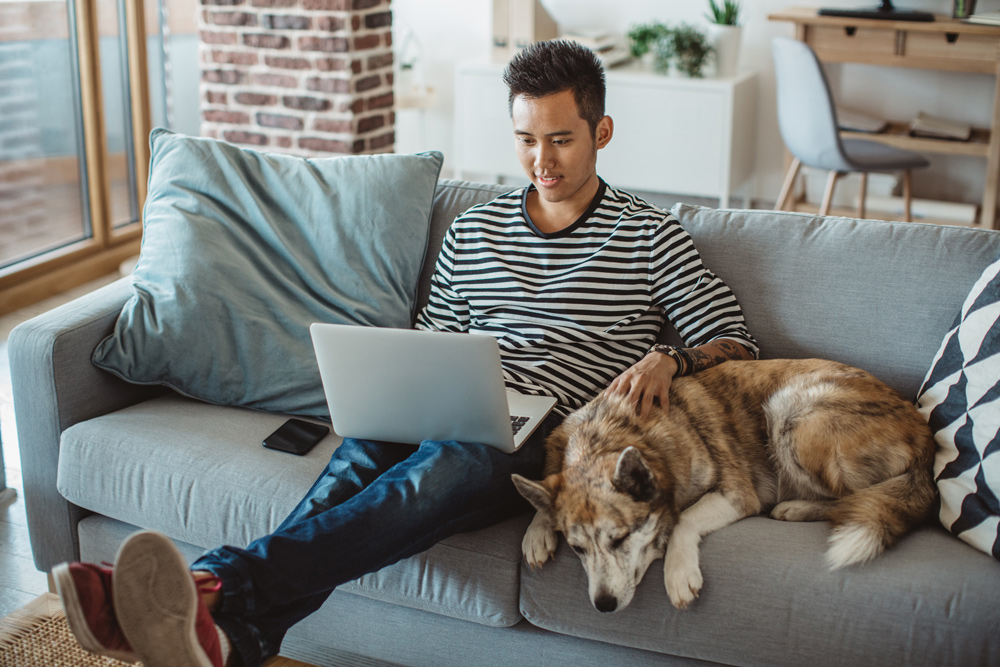 man sits on couch with dog while investing with laptop